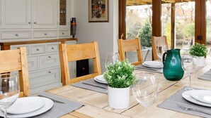 A dining area with a wooden table set for four, surrounded by white cabinets and a large window with a view of greenery. - Serene Riverside 6BR Luxury Retreat in Curdridge (Southampton)