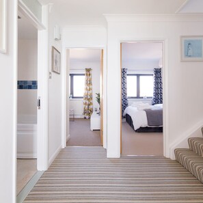 A hallway leading to a bedroom with a bed, a bedside table, and a window with curtains. - The Sea House - Ultimate Seafront Living Sunsets (Chichester)