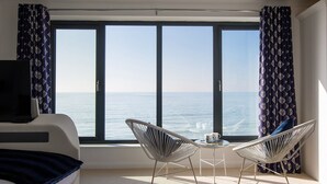 Ein Zimmer mit großem Fenster mit Meerblick, zwei weiße Stühle, ein kleiner Tisch mit einer Tasse und ein Bett mit einer dunkelblauen Decke.