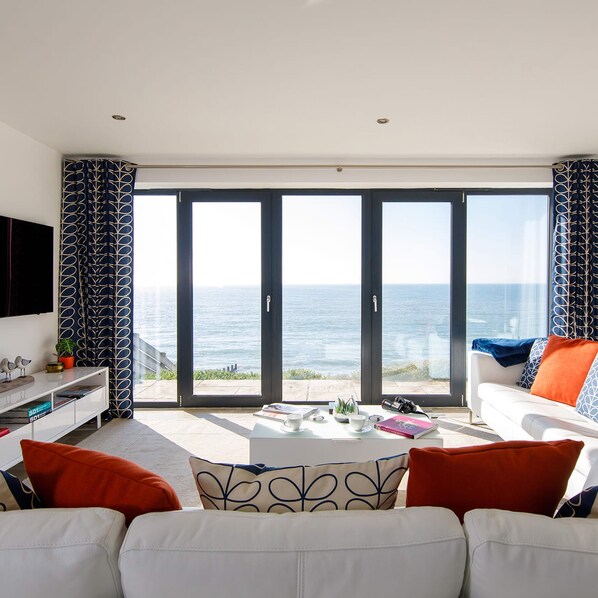 A modern living room with a white sofa, a coffee table, and a view of the sea through large glass doors. - The Sea House - Ultimate Seafront Living Sunsets (Chichester)
