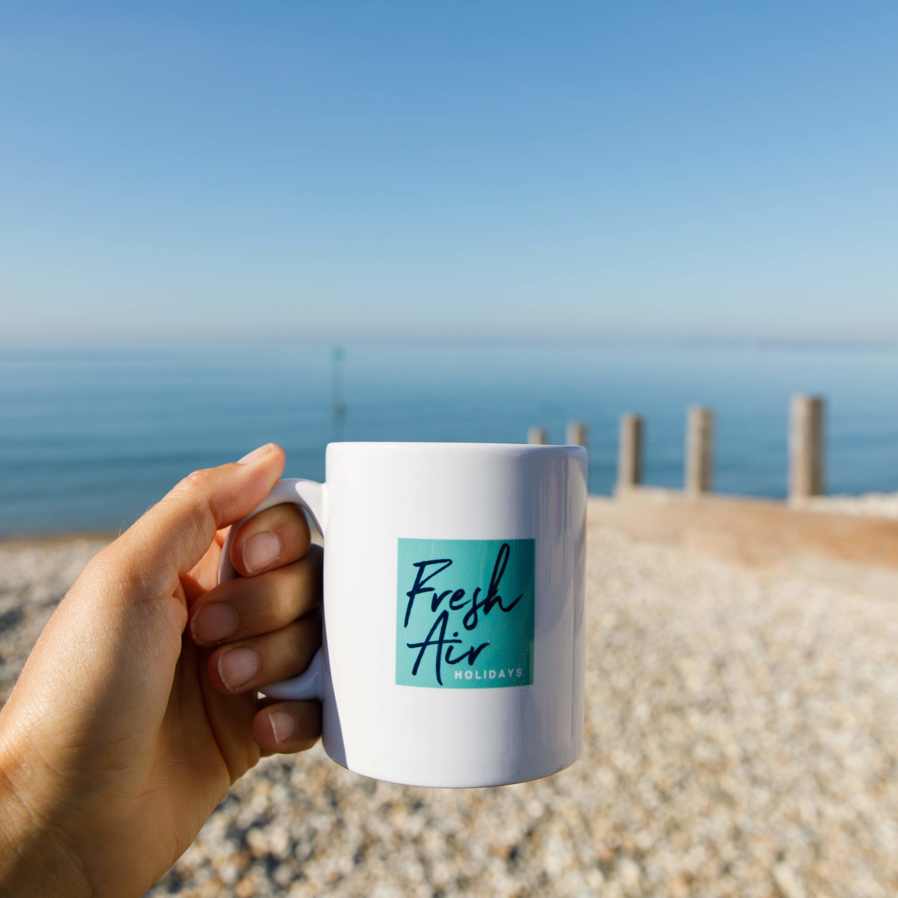 A hand holding a white mug with "Fresh Air" and "HOLIDAYS" text on a beach.