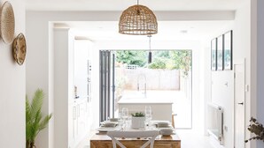 Une cuisine lumineuse et un espace repas à aire ouverte, avec une table en bois dressée pour deux personnes, des chaises blanches et un luminaire suspendu en fibres tressées.