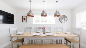 A dining area with a wooden table set for six, white chairs, and three pendant lights. - Beach House for 10 With hot tub Garden (Chichester)