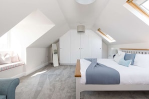 Ein Schlafzimmer mit schrägen Wänden, einem Bett mit blauer Bettwäsche, einem Nachttisch, einem Sessel und einem Kleiderschrank.