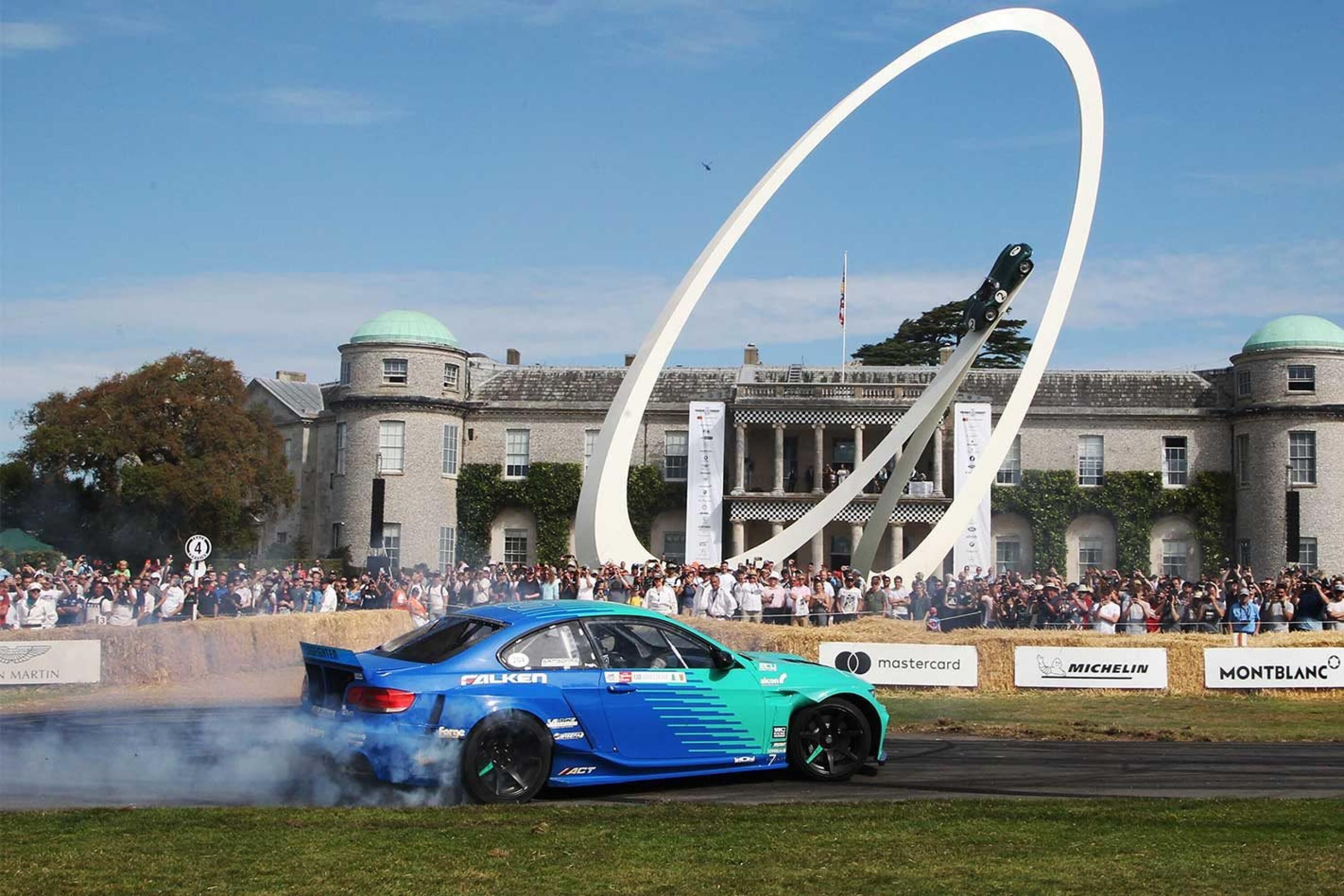A blue rally car with smoke, a crowd, and a building with a large white arch in the background.