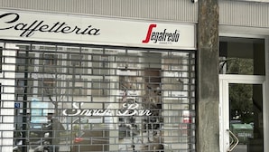 Un escaparate con una persiana metálica, un cartel que dice "Caffetteria Segafredo Zanetti" y el reflejo de un coche y árboles en el cristal.