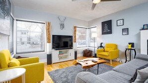 Un salon moderne avec un canapé gris, des fauteuils jaunes, une table basse en bois et un téléviseur à écran plat.