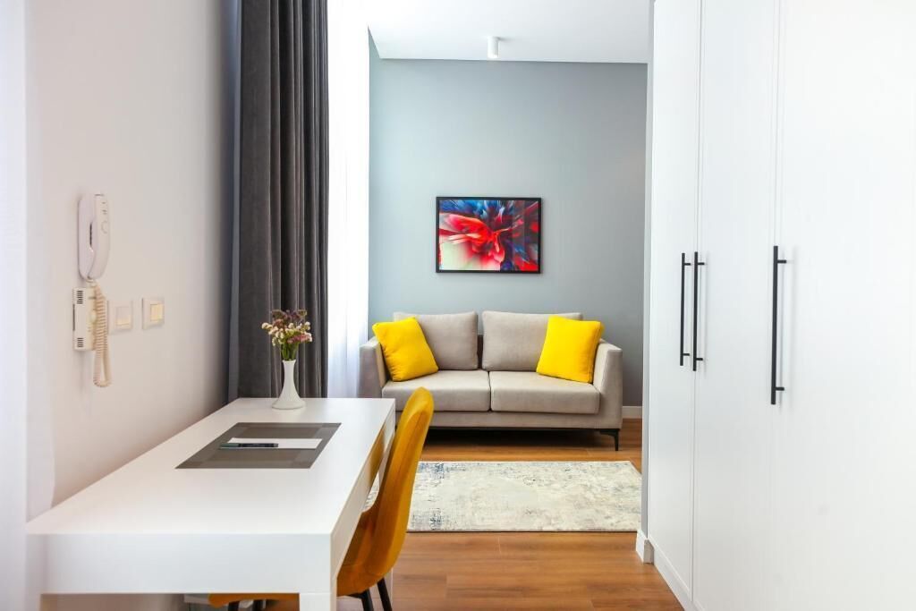 Una sala de estar moderna con un sofá blanco, cojines amarillos, un cuadro abstracto gris y un comedor con una mesa blanca y un jarrón de flores.