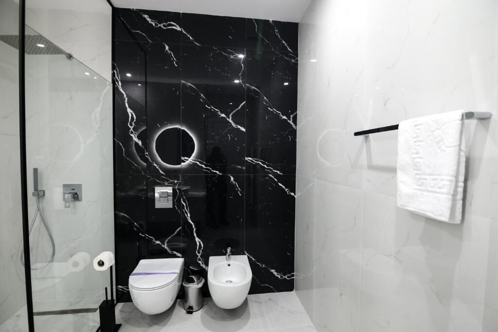 A modern bathroom with a black marble wall, a glass shower enclosure, a white toilet, a bidet, and a towel rack with a white towel.