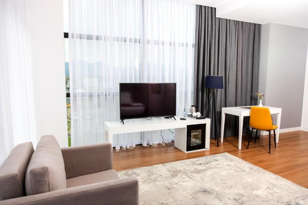 Ein modernes Wohnzimmer mit einem Flachbildfernseher auf einem weißen Konsolentisch, einer Couch, einem kleinen Tisch mit Lampe und einem Sessel.