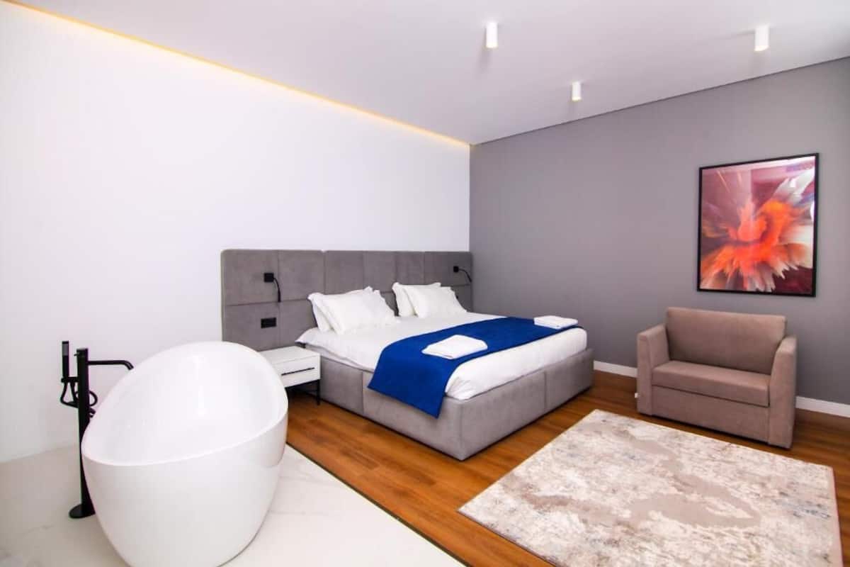 A modern bedroom with a large bed, a white bathtub, a grey sofa, and a large painting on the wall.