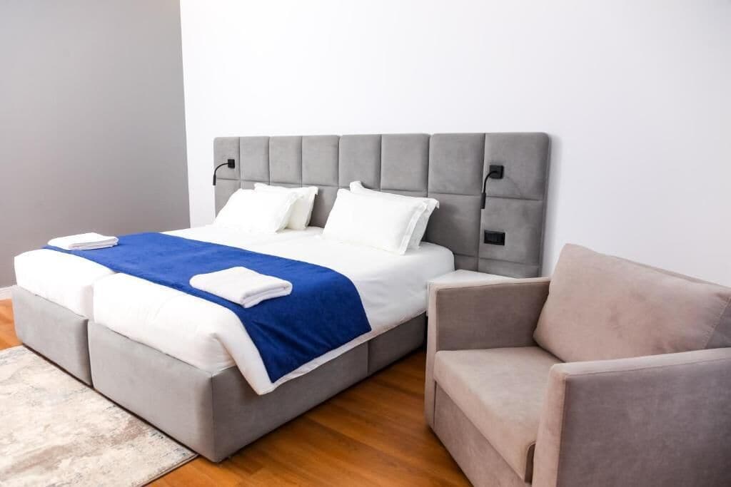 Un dormitorio moderno con una cama tapizada de gris, ropa de cama blanca y azul, y un sillón tapizado de gris.