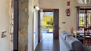 A modern interior with a stone wall, a grey sofa, a dining area with a table and chairs, and a view of a swimming pool and mountains. - Finca Macías (Cártama)