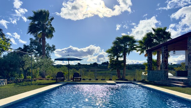 A swimming pool with two chairs under an umbrella, surrounded by palm trees and a stone building. - Finca Macías (Cártama)