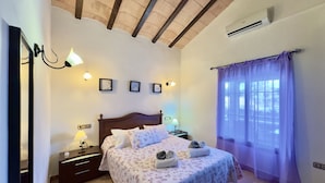 A bedroom with a wooden ceiling, a bed with floral bedding, a nightstand with a lamp, and a window with purple curtains. - Finca Macías (Cártama)