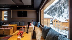 A modern interior with a wooden bench seating area, a flat-screen TV mounted on the wall, and a view of snow-covered buildings through large windows.