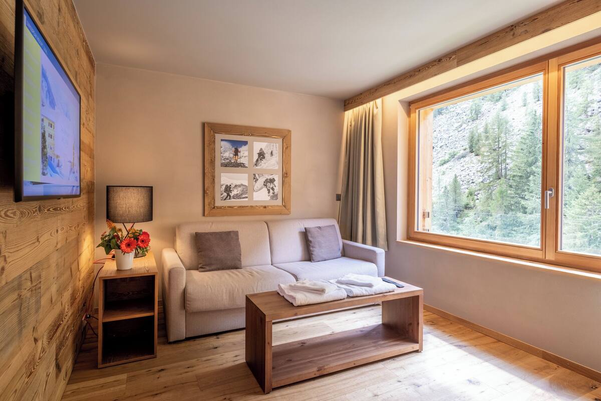 A cozy room with a wooden floor, a sofa, a coffee table, a TV, a framed picture, and a window with a forest view.