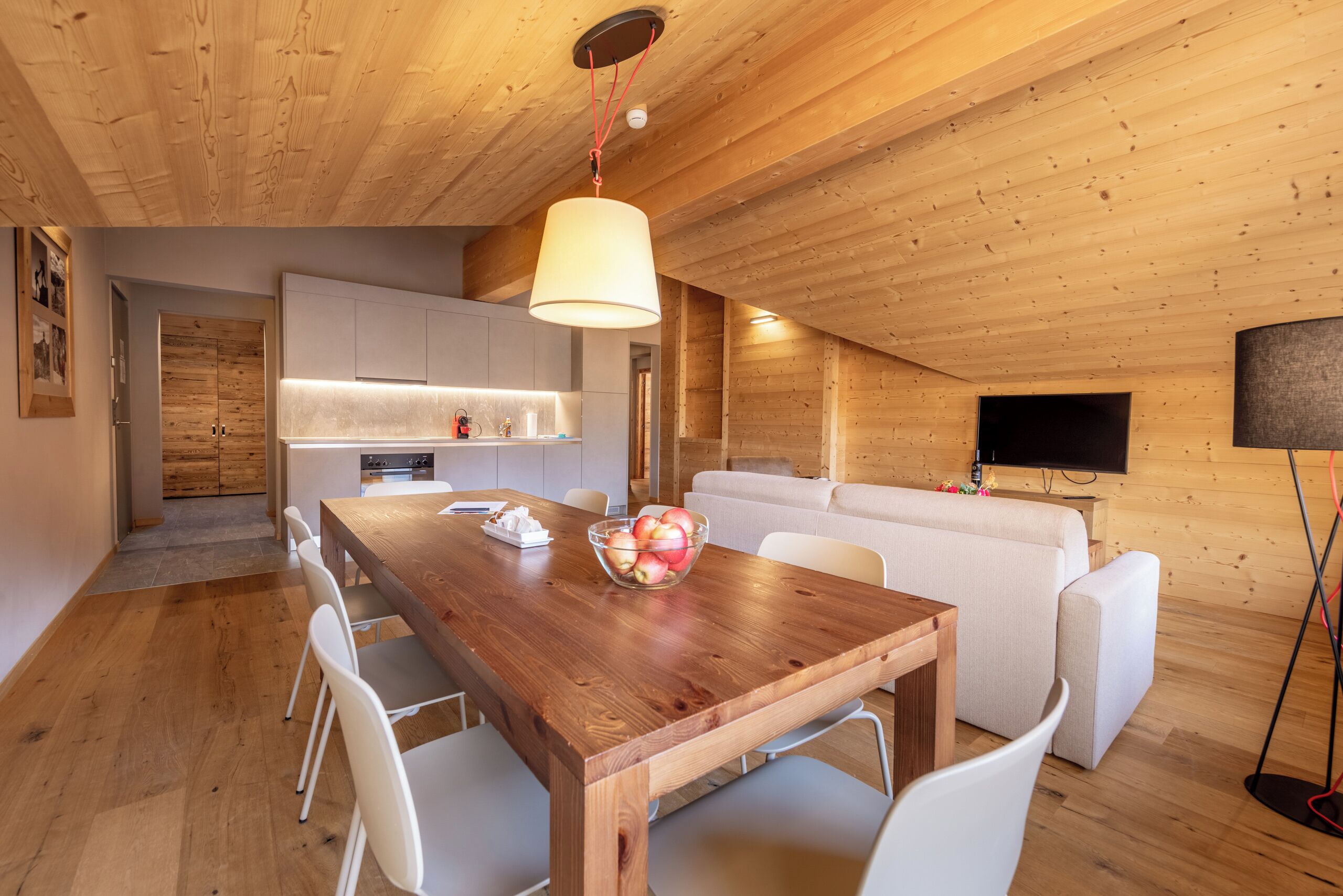 A wooden interior with a dining table, white chairs, a sofa, and a TV mounted on the wall.