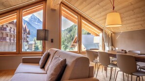A modern interior with a sofa, dining table, chairs, and large windows offering mountain views.