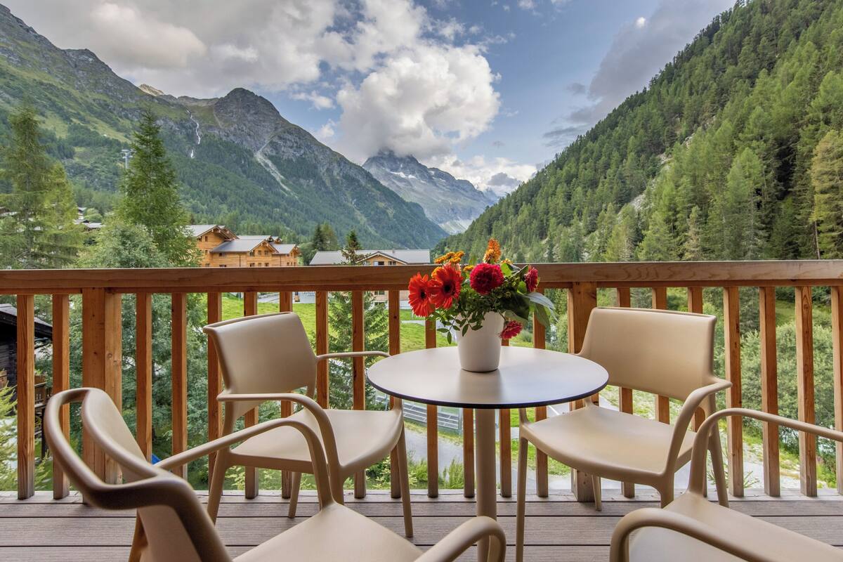 A wooden deck with chairs and a table, overlooking a mountainous landscape with houses in the valley.