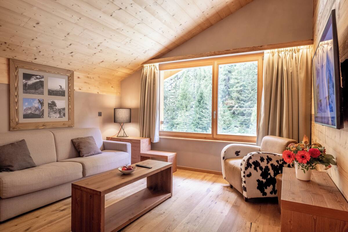 A cozy living room with a wooden ceiling, a sofa, a coffee table, a chair, a TV, and a window with curtains.