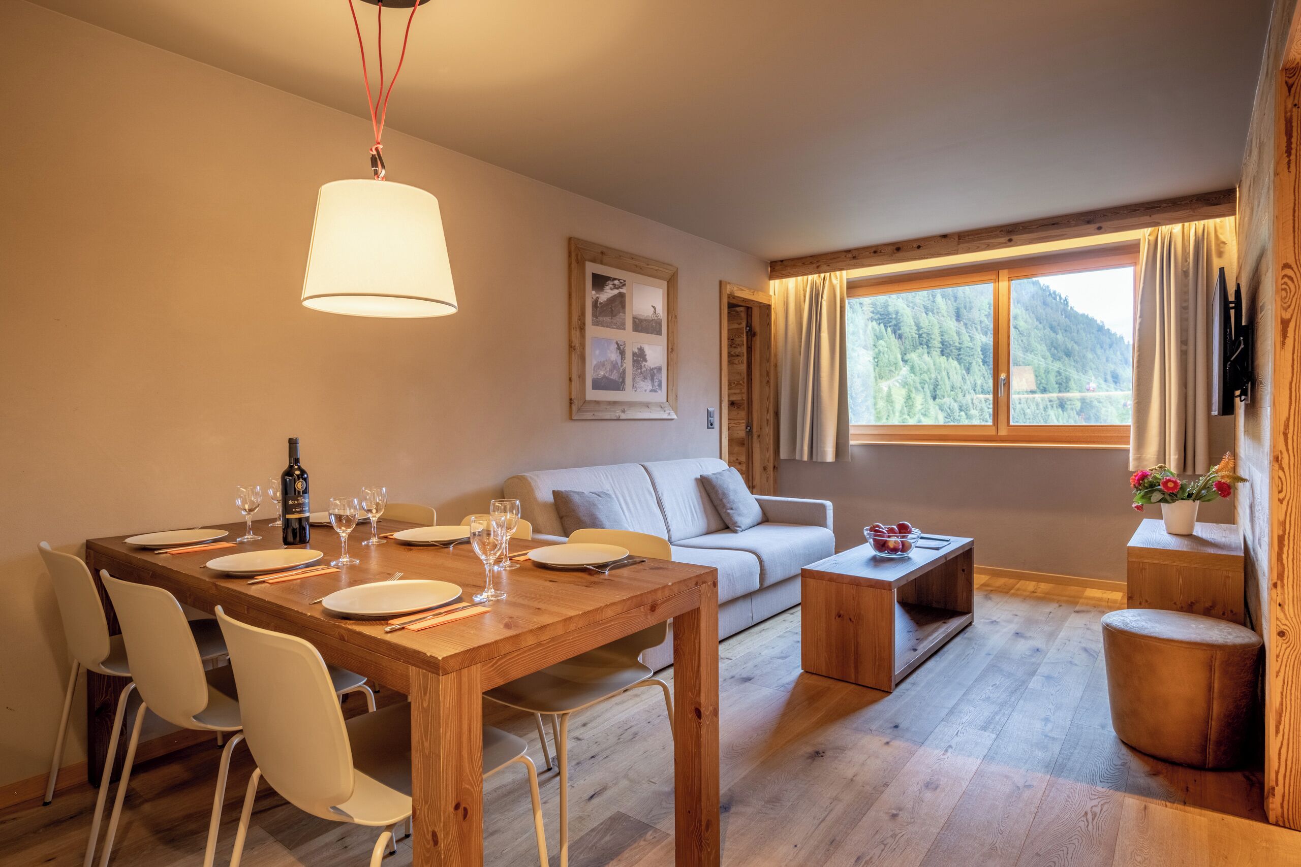 Ein Essbereich mit einem Holztisch, weißen Stühlen, einer Couch und einem Fenster mit Blick auf einen Wald.