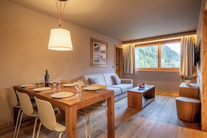 A dining area with a wooden table, white chairs, a sofa, and a window with a view of a forest. - SWISSPEAK Resorts by Pierre & Vacances Premium Zinal (Anniviers)