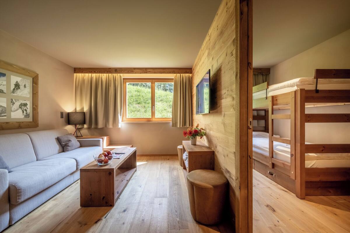 A cozy room with a sofa, wooden coffee table, a small side table with a vase, a bunk bed, and a window with curtains.