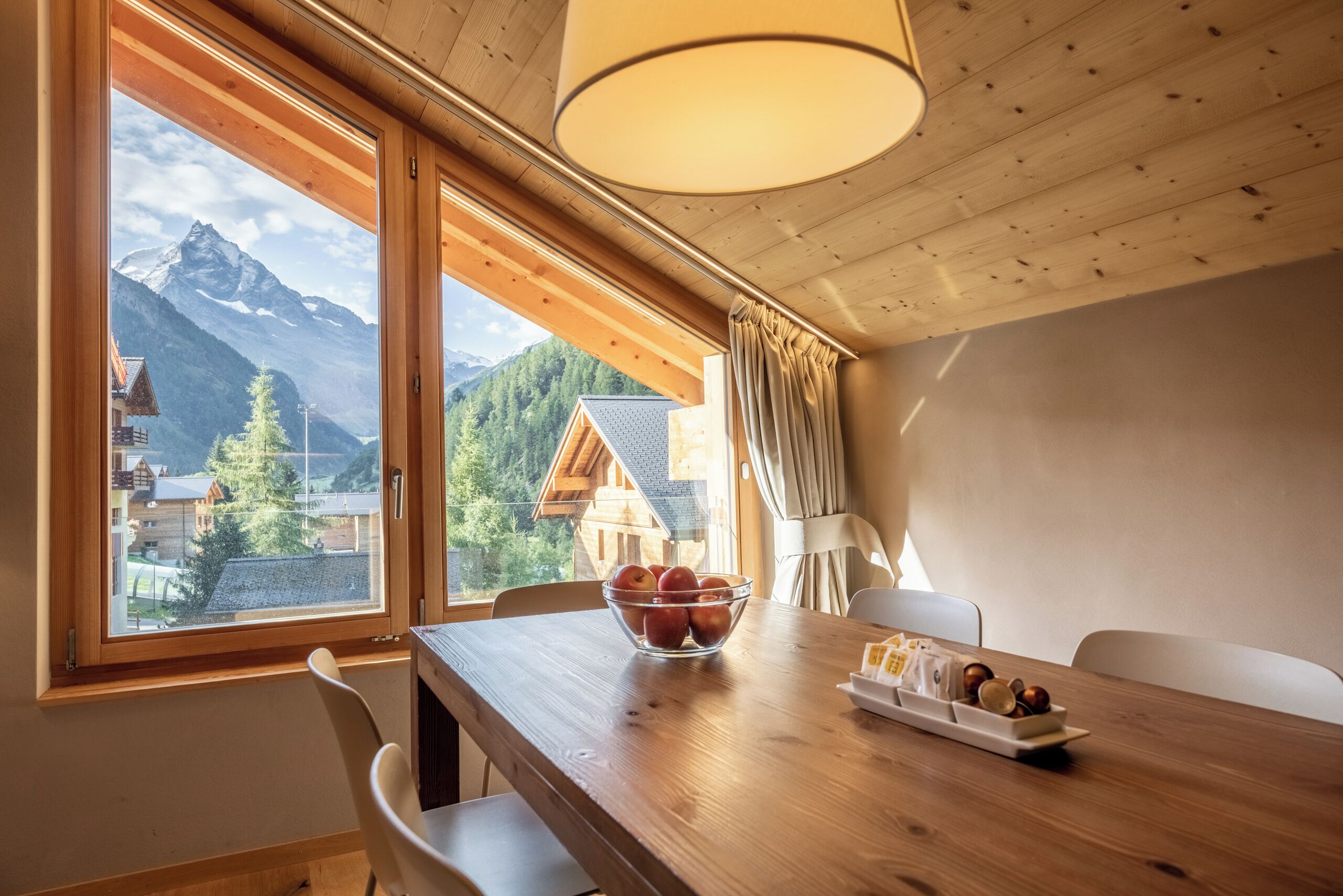 Una mesa de comedor de madera con un bol de manzanas y una bandeja de nueces, una ventana con vistas a la montaña y un techo de madera con una lámpara colgante.