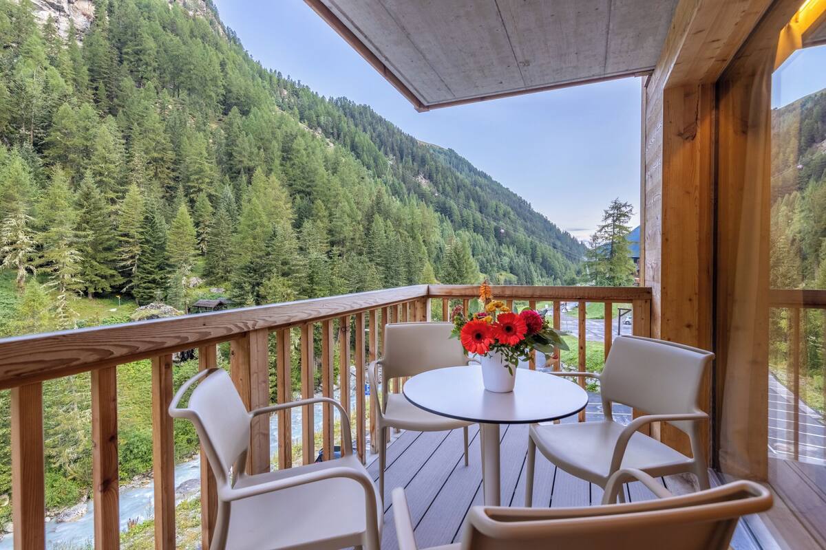 A wooden deck with a table and chairs, overlooking a river and forested mountains.
