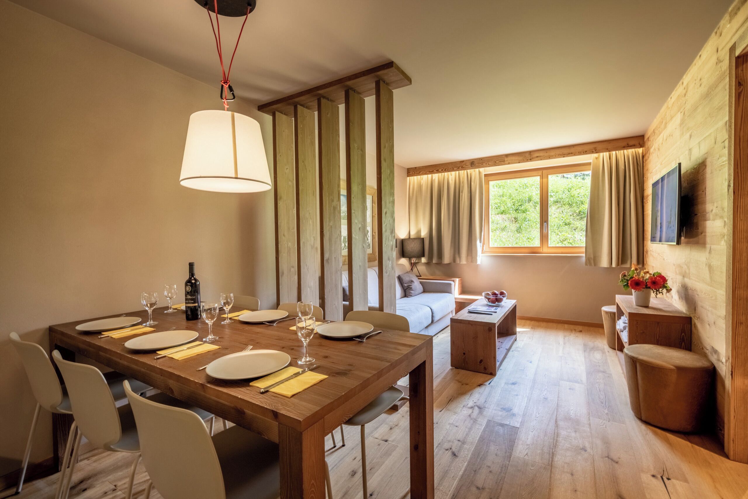 A dining area with a wooden table set for six, a sofa, a TV mounted on the wall, and a window with curtains.