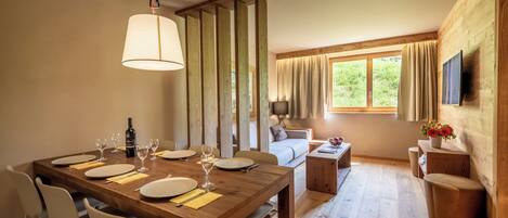 A dining area with a wooden table set for six, a sofa, a TV mounted on the wall, and a window with curtains.