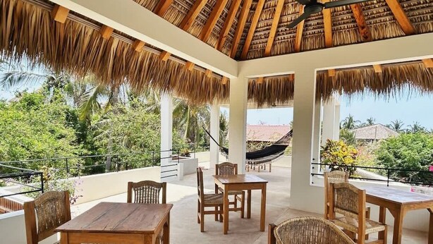 An outdoor patio with wooden furniture, a hammock, and a view of a red-roofed building. - Casa Aymara 601: Beach, Starlink & Privacy (Brisas de Zicatela)