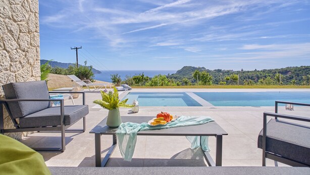 Zona de piscina con sillones, una mesa con una bandeja de frutas y vistas al mar y a las colinas.