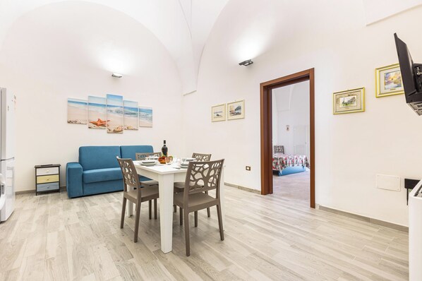 Una sala de estar moderna con un sofá azul, una mesa de comedor con sillas y una zona de cocina con refrigerador y un mueble pequeño.