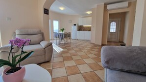 A living room with a beige sofa, a grey sofa, and a pink potted orchid. There is a kitchen in the background with white cabinets and a tiled floor. - Nice apartment near beach and city ​​centre (Crikvenica)