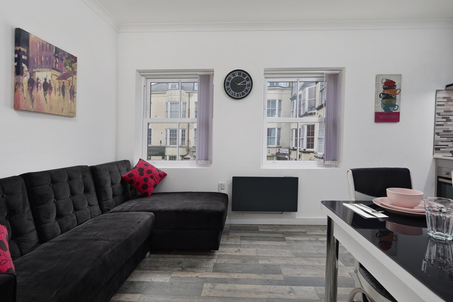 Una sala de estar moderna con un sofá modular negro, una mesa de comedor con platos rosas y dos ventanas con vistas a edificios.
