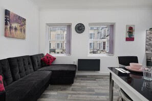 A modern living room with a black sectional sofa, a dining table with pink plates, and two windows with a view of buildings. - 1 Bed Flat - Sleeps 2 - Wifi (Scarborough)