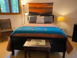 A bedroom with a bed, a nightstand, a lamp, a chair, and a window with a view of trees. - Beaver Dome Geodesic Lake Home 3br 2bath (Rogers)