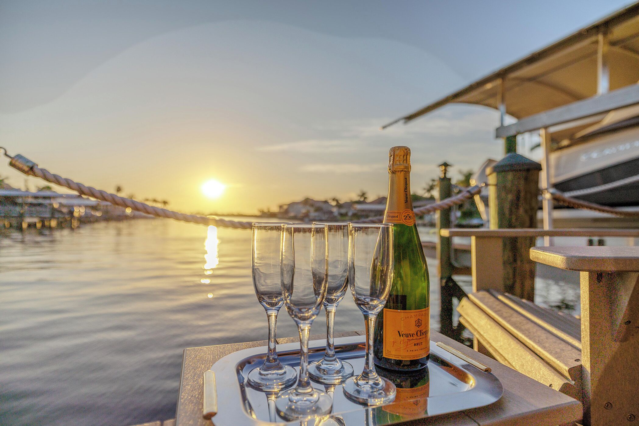 A bottle of Veuve Clicquot champagne and four glasses on a tray by a waterfront.