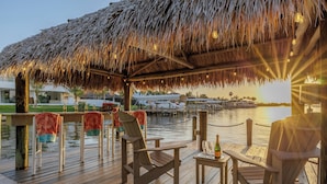 A wooden deck with chairs and a table, overlooking a body of water with a thatched roof overhead.