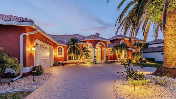 A residential property with a paved driveway, a garage, and a well-maintained garden with palm trees and shrubs.