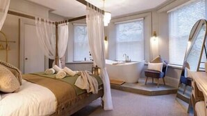 A four-poster bed with a canopy, a sitting area with a chair, a bathtub, and a large mirror.