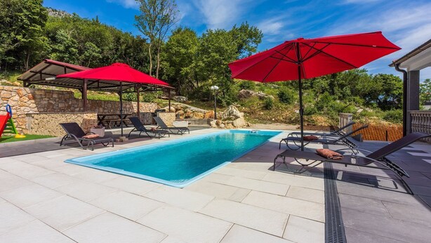 A swimming pool with lounge chairs and red umbrellas, surrounded by stone walls and greenery.