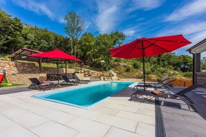 A swimming pool with lounge chairs and red umbrellas, surrounded by stone walls and greenery.