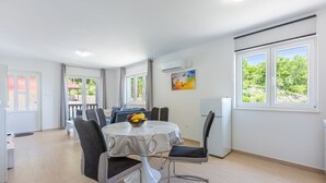 A modern, well-lit dining area with a round table, chairs, and a fruit bowl. There is a window with a view of greenery outside.