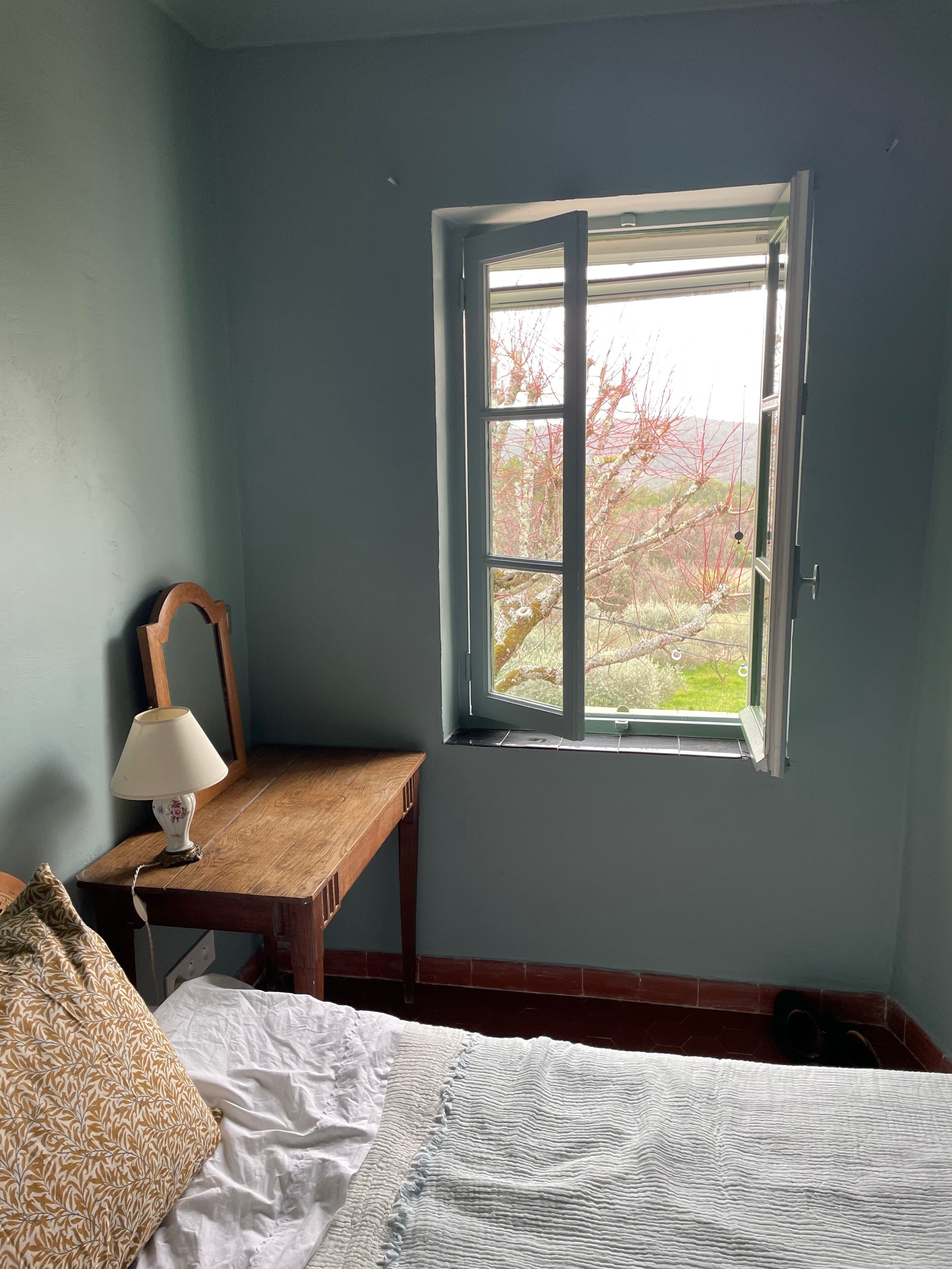 A bedroom with a wooden desk, a lamp, a bed, and a window with a view of trees.