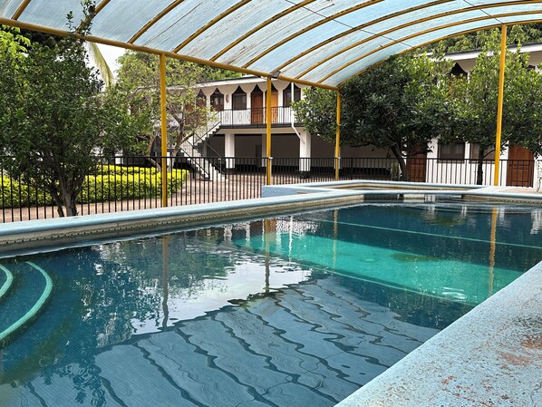 A swimming pool with a curved edge, surrounded by a covered area with yellow pillars and a white building in the background.
