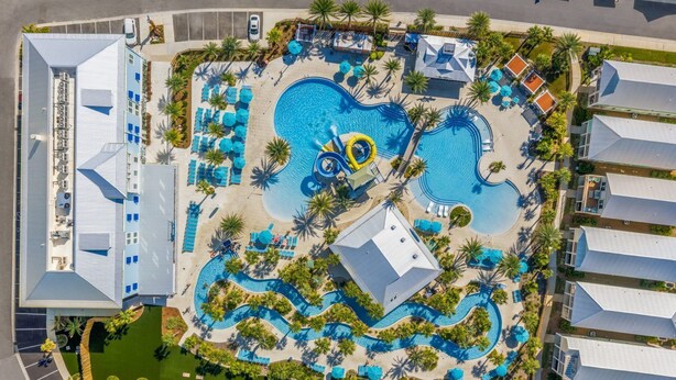 An aerial view of a resort with a central swimming pool, multiple lounge areas, and palm trees.
