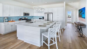 A modern kitchen with white cabinetry, a central island with bar stools, a stainless steel refrigerator, and a view of the beach through the door.
